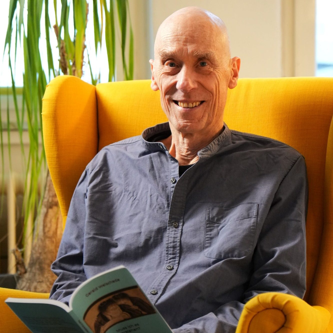 Gerald Weischede sitzt lächelnd in einem gelben Sessel und hält ein Buch in der Hand.