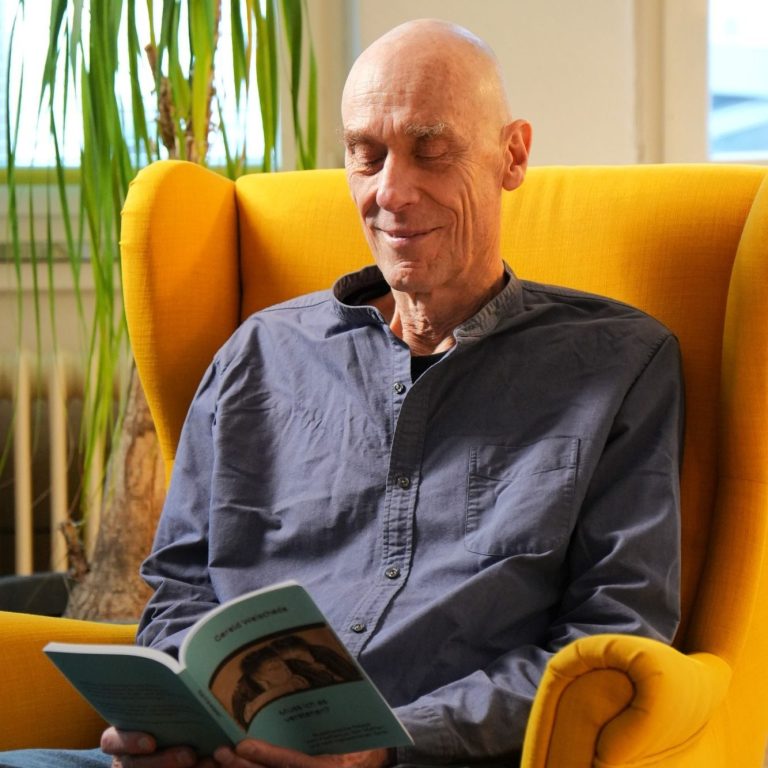 Gerald Weischede sitzt lächelnd in einem gelben Sessel und liest ein Buch. 